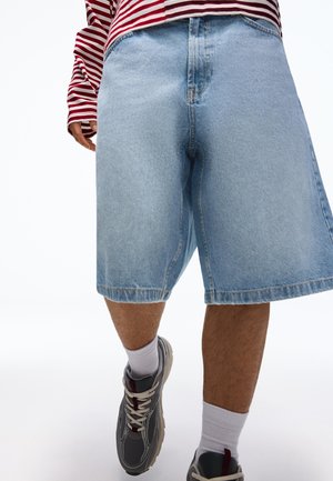 Person trägt hellblaue Jeansshorts, ein rot-weiß gestreiftes Langarmshirt, weiße Socken und graue Turnschuhe und geht vorwärts.