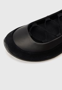 Chaussure plate de ballet noire avec un bout arrondi, des brides élastiques croisées et une semelle texturée sur fond clair.