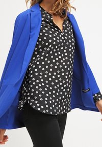Blazer blu sopra una blusa nera con pois bianchi, dotata di un colletto con bottoni e tessuto fluido. Pantaloni neri completano l'outfit.