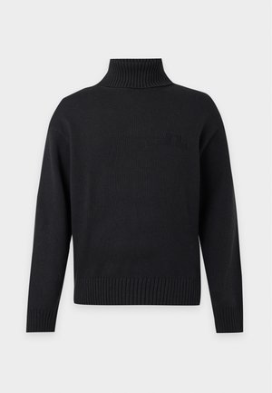 Joven con un jersey negro de cuello alto, pantalones oscuros y zapatillas negras de pie frente a un fondo blanco liso.
