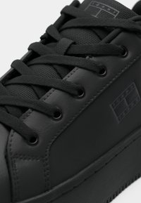 Černá teniska z hladké kůže s texturovaným látkovým jazykem, vybavená černými tkaničkami, průchodkami a logem na boční straně.