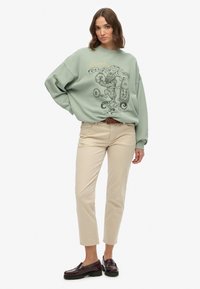 Mintgroene sweatshirt met tijgerafbeelding; ruime pasvorm. Beige broek en bordeauxrode loafers. Casual outfit met minimalistisch ontwerp.