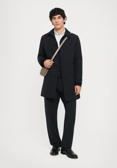Marineblauer Mantel mit spitzem Kragen, Knopfverschluss und großen Taschen, getragen über einem dunklen Pullover und Hose, kombiniert mit schwarzen Schuhen.