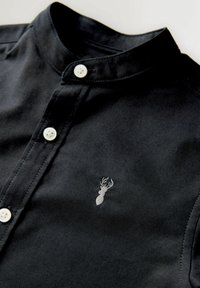 Maglia nera con collo alto, caratterizzata da bottoni bianchi e un sottile emblema grigio di cervo sul petto a sinistra. Tessuto dalla texture liscia.