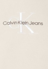 Tessuto in pile beige con logo ricamato "Calvin Klein Jeans" in marrone scuro, caratterizzato da un prominente monogramma bianco "CK".