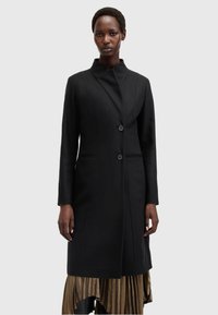 Cappotto di lana nera con colletto alto, dal design essenziale, con due bottoni e tasche laterali. Indossato sopra una gonna dorata a pieghe.
