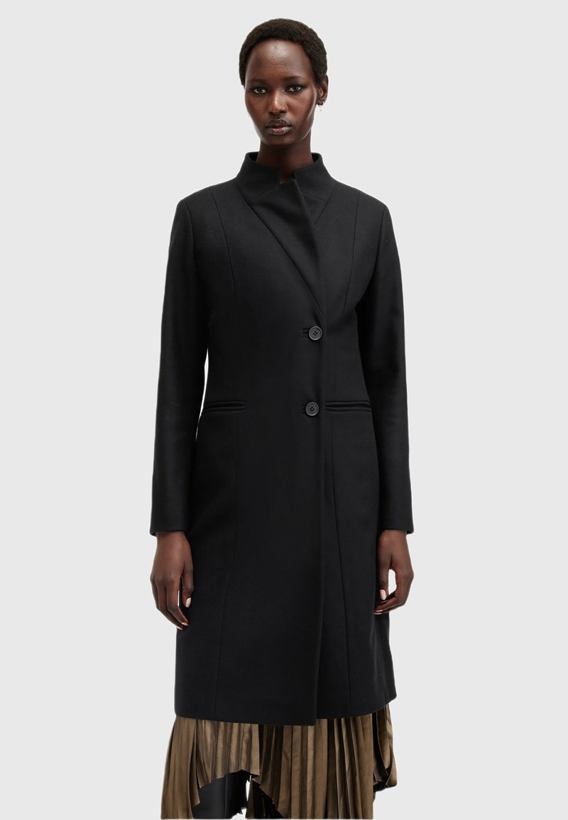 Cappotto di lana nera con colletto alto, dal design essenziale, con due bottoni e tasche laterali. Indossato sopra una gonna dorata a pieghe.