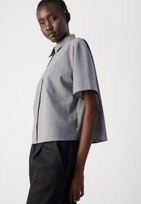 Short-sleeve, light gray shirt with a collar, featuring a single chest pocket and a boxy shape. Smooth fabric with side slits for comfort.