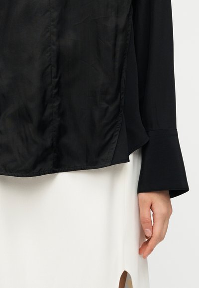 Μπλούζα από μαύρο σατέν ύφασμα με χαλαρή εφαρμογή, φουσκωτά μανίκια και στρογγυλεμένη ραφή στο τελείωμα, συνδυασμένη με κρεμ φούστα με σκίσιμο στο πλάι.