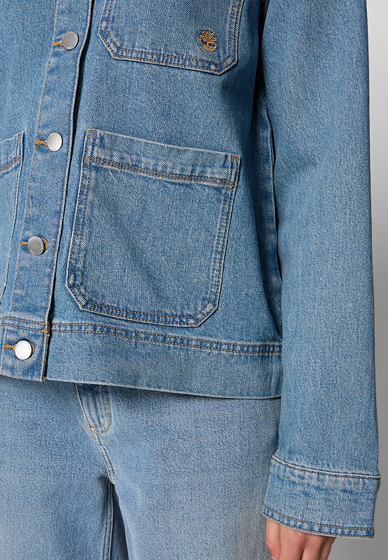 Giubbotto in denim azzurro chiaro con bottoni argento, due tasche frontali e dettagli di cucitura visibili. Progettato con una vestibilità rilassata e maniche lunghe.