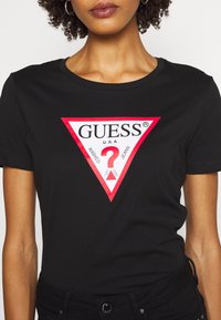 Kvinde iført sort tætsiddende t-shirt med rødt og hvidt Guess-logo i form af en trekant, der har teksten "Guess U.S.A." og et rødt spørgsmålstegn.