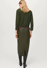 Maglione verde a maglia con polsini arrotolati, abbinato a una gonna midi in pelle verde scuro con spacco posteriore e accenti bordeaux. Stivali neri fino al ginocchio.