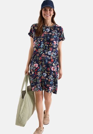 Femme portant une robe fleurie bleu marine, une casquette bleu marine, des chaussures beige faciles à enfiler, portant un grand sac cabas matelassé vert clair, souriante et marchant vers l'avant.