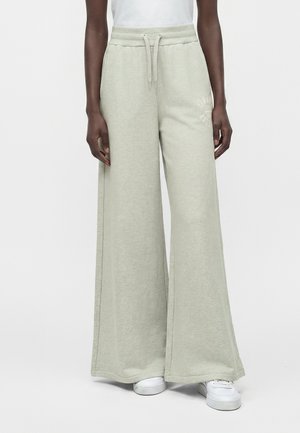 Personne porte un jogging ample gris clair avec une taille à cordon et des baskets blanches sur un fond clair uni.