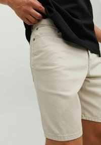 Shorts in cotone beige con tasche laterali, chiusura frontale con bottone e cuciture a contrasto. Modello che indossa una maglietta nera, mano sulla vita.