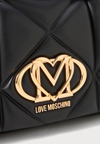 Mustra musträhine ehetega must kunstnahast kott, mille esiküljel on silmapaistev kullavärvi logo, kus on kokku põimitud tähed "M" ja "W".