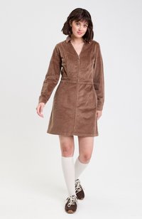 Robe en velours côtelé marron avec des manches longues, un col et une fermeture éclair à l'avant. Le mannequin l'a associée à des chaussettes blanches jusqu'au genou et à des chaussures brunes à lacets.