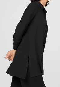 Mustapaita, jossa on pitkä hiha ja terävä kaulus, suorassa leikkauksessa, sivuhalkiot ja sileä rakenne. Ei näkyviä kuvioita tai varusteita.