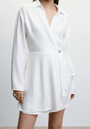 Mujer vistiendo un vestido envolvente de manga larga blanca texturizada con un cuello y un broche plateado en la cintura.