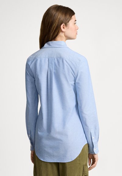 Γυναίκα με μακριά καστανά μαλλιά, φορώντας ανοιχτό μπλε πουκάμισο με μακριά μανίκια και παντελόνι ελιάς, γυρισμένη πλάτη μπροστά σε απλό φόντο.