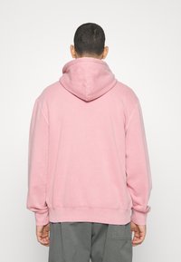 Růžová mikina s kapucí a volným střihem. Má žebrované manžety a lem, měkký povrch a velkou kapuci vzadu.