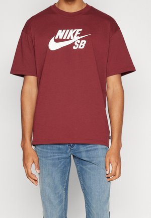 Person, die ein weinrotes Nike SB T-Shirt und hellblaue Jeans trägt, steht mit entspannten Armen an den Seiten vor einem einfarbigen Hintergrund.