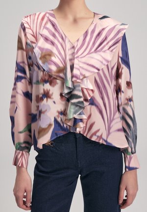 Femme portant un chemisier pastel à fleurs et rayures avec un devant à volants et des manches longues, associé à un jean foncé.