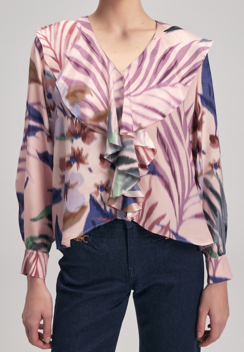 Femme portant un chemisier pastel à fleurs et rayures avec un devant à volants et des manches longues, associé à un jean foncé.