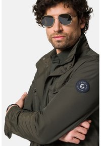 Oliwkowa kurtka z kołnierzem, z zatrzaskami i kieszeniami z przodu. Posiada naszywkę z logo na rękawie i jest noszona z srebrnymi okularami przeciwsłonecznymi.