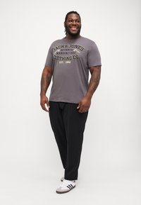 Pelēka t-krekls ar "JACK & JONES" logo dzeltenā krāsā, apvienots ar melniem biksēm un baltām apaviem, kas rotāti ar melnām un brūnām svītrām.