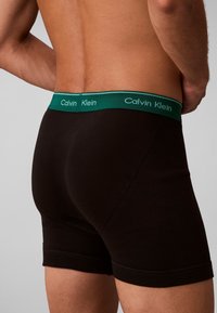 Muž v čiernych boxerkách so zeleným pásom s nápisom Calvin Klein, zobrazený zospodu do polovice stehna.