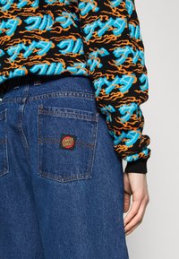 Sweater de malha colorido com padrões em preto, azul e laranja, combinado com calças de ganga azul-escuro apresentando um rótulo vermelho e preto 'Santa Cruz' no bolso.