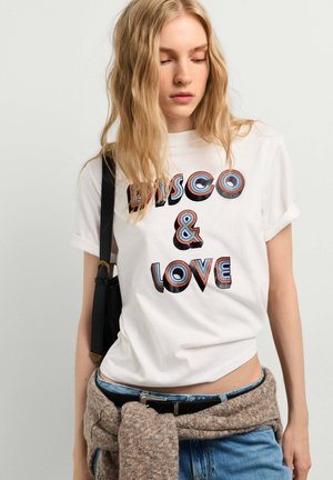 Giovane donna con lunghi capelli biondi indossa una maglietta bianca con la scritta "DISCO & LOVE,", jeans blu e un maglione marrone legato in vita.