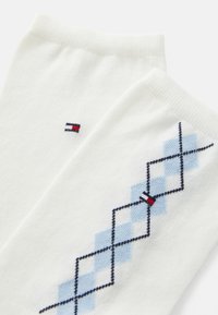 Vita bomullssockor med ett blått och svart argyle-mönster, med en liten röd, vit och blå logodetalj nära kanten.