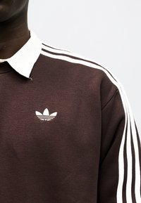 Személy barna Adidas pulóvert visel, fehér háromcsíkos mintával az ujjain és fehér gallérral, egy egyszínű háttér előtt.