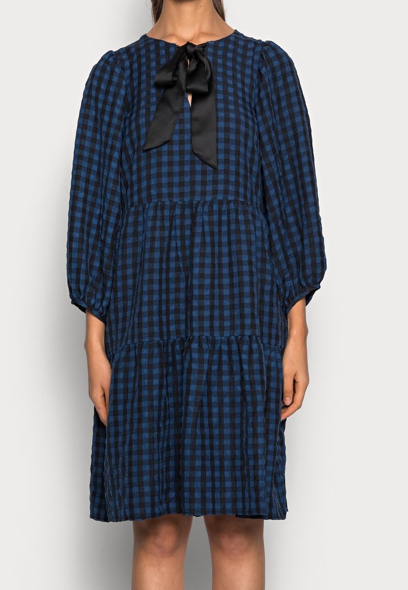 Robe à carreaux bleus et noirs avec un décolleté rond, de longues manches bouffantes et un nœud en satin noir à l'avant. Présente un ourlet à volants.