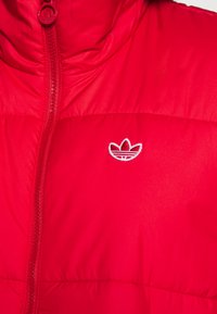 Röd puffjacka med hög krage, försedd med en central dragkedja och en broderad logotyp i vitt på vänster bröst.