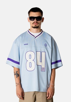 Mand iført lys blå oversized trøje med nummer 84, lilla striber på ærmerne, solbriller og beige bukser mod en ensfarvet baggrund.