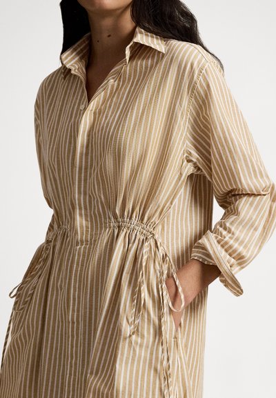 Femme portant une robe à manches longues beige et blanche à rayures verticales, taille cintrée et poches latérales, main posée dans la poche droite.