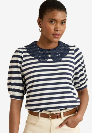T-shirt à rayures bleu marine et blanches à manches courtes, avec un col en dentelle texturé, associé à une ceinture marron sur un pantalon crème.