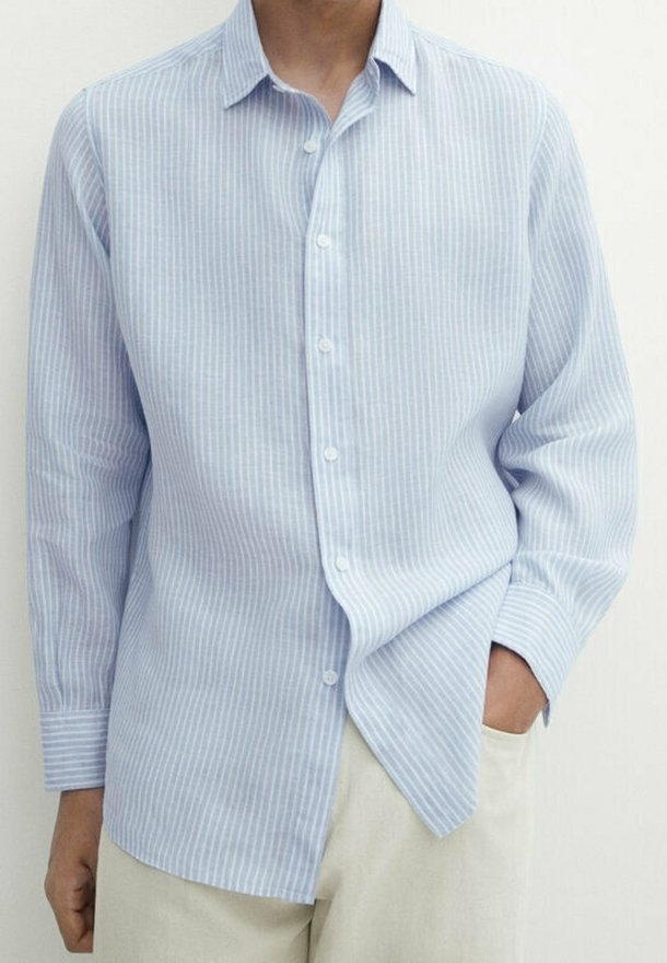 Camicia azzurra a righe con bottoni, con maniche lunghe, realizzata in un tessuto intrecciato. Presenta un colletto e un orlo leggermente curvato. Abbinata a pantaloni chiari.