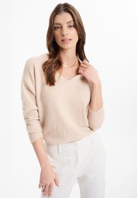 Pullover beige lavorato a maglia con scollo a V, spalle basse, polsini a coste e una vestibilità rilassata, abbinato a pantaloni bianchi su misura.