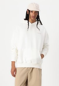 Sudadera blanca con capucha y bolsillo tipo canguro, que presenta un sutil logo en color beige claro. Combinada con pantalones beige claros.