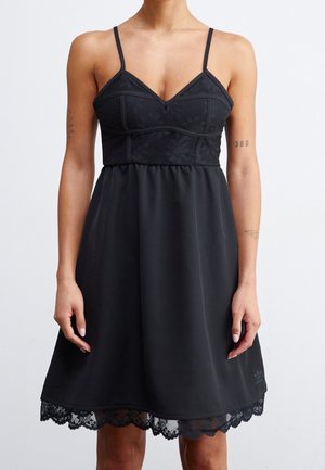 Femme portant une robe noire sans manches avec des détails en dentelle sur le buste et l'ourlet, debout, les bras détendus le long du corps, sur un fond blanc uni.