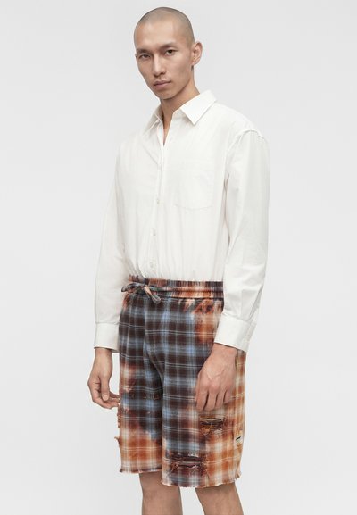 Άνδρας που φοράει λευκό πουκάμισο με κουμπιά, μαζεμένο μέσα σε πολύχρωμο καρό σορτς με σκισίματα και ζωνάκι με κορδόνι, στέκεται μπροστά σε απλό φόντο.