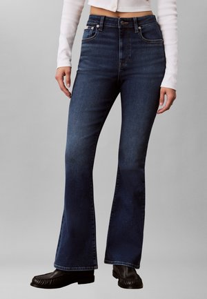 Persona che indossa jeans a vita alta svasati blu scuro, scarpe nere e un top bianco corto a maniche lunghe su sfondo semplice.
