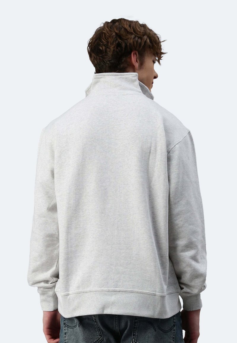 Uomo che indossa una felpa grigio chiaro con collo alto, visto da dietro, con le mani rilassate ai lati e jeans scuri visibili.