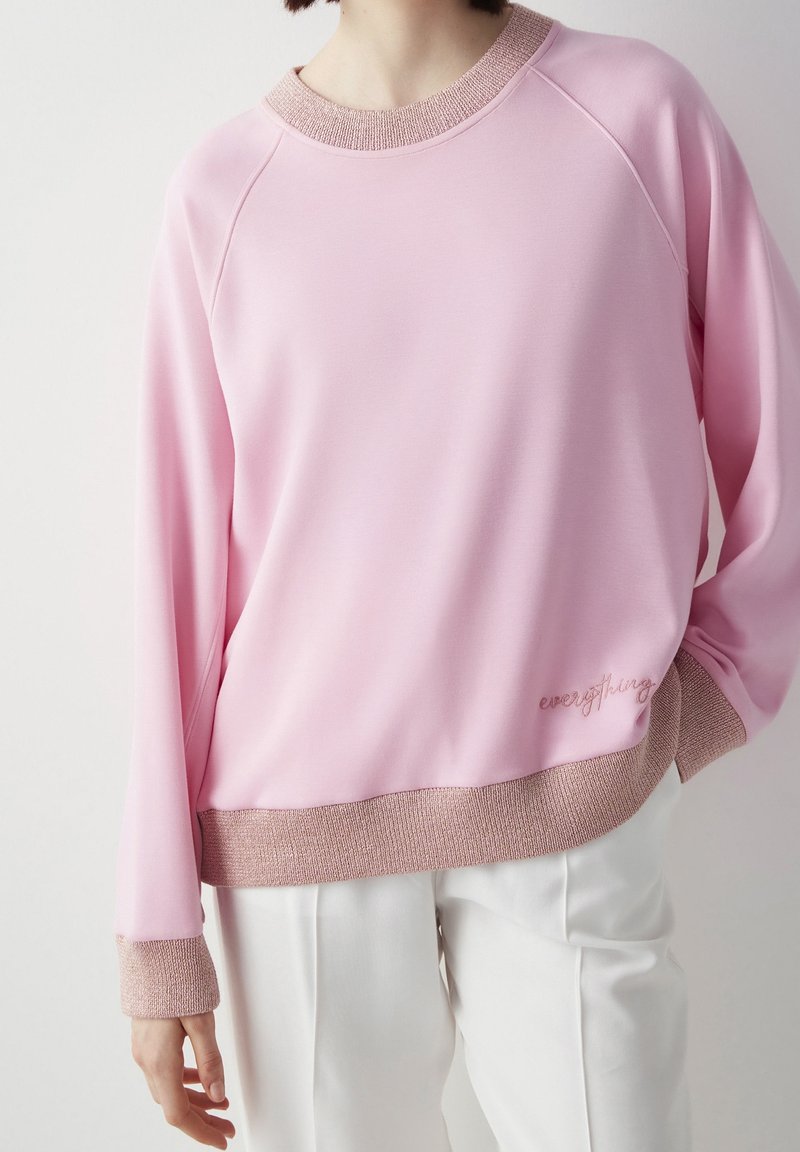 Personne portant un pull rose clair avec des poignets et un ourlet côtelés, un pantalon blanc et une posture détendue avec une main dans la poche.