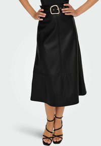 Femme portant une jupe midi noire en simili cuir avec une ceinture à boucle dorée et des sandales à talons hauts à lanières noires, les mains sur les hanches.