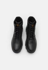 Botas de cuero negro con puntera redonda, diseño resistente de cordones, cuello acolchado y suela de goma texturizada. Presenta un logo dorado en la lengüeta.